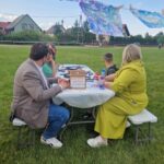 pQ0QUheC Na zdjęciu grupa osób siedzących przy białym stole na zielonej trawie. Nad nimi wiszą kolorowe obrazy na sznurkach. W tle widać domy i niebo z chmurami.