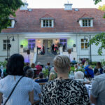 Na zdjęciu widać koncert na tle białego budynku z czerwonym dachem. Przed sceną siedzi publiczność w ogrodzie. W tle widać zielone drzewa.