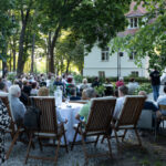 Grupa ludzi siedzi na zewnątrz na krzesłach i stołach pod drzewami. Tło to biały budynek z czerwonym dachem. Scena jest zielona i oświetlona naturalnym światłem.