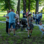 Na zdjęciu grupa ludzi w parku, niektórzy z wózkiem, inni siedzą. Tło zielone, drzewa i trawa. Letnie stroje, jasne kolory.