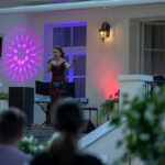 Kobieta w eleganckiej sukni śpiewa na scenie z mikrofonem. Tło w fioletowych i czerwonych światłach. Obok klawiatura i dekoracje z zielenią.