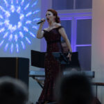 Piosenkarka w ciemnej, połyskującej sukni śpiewa na scenie. Tło oświetlone kolorowymi światłami i różowym odcieniem. W tle klawiszowiec i scena z roślinami.