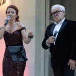 Na zdjęciu widać kobietę w czerwonej sukni i mężczyznę w czarnym garniturze, oboje z mikrofonami. Tło jest jasne, z latarnią na ścianie. Kobieta śpiewa, mężczyzna stoi obok.