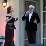 Dwie osoby śpiewają na scenie. Kobieta w czerwonej sukni, mężczyzna w ciemnym garniturze. Tło jasne, z delikatnym oświetleniem.