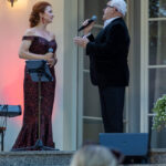 Dwoje ludzi śpiewa na scenie, kobieta w czerwonej sukni, mężczyzna w czarnym garniturze. Tło jasne z lampą.