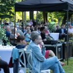 Na zdjęciu widać zgromadzenie ludzi na otwartym terenie, siedzących przy białych stołach pod czarnym namiotem, w otoczeniu zieleni. W tle widoczne są drzewa, a atmosfera wydaje się być spokojna i przyjazna. Dekoracje stołów