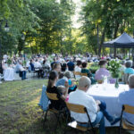 Spotkanie plenerowe w parku, ludzie siedzą przy białych stołach, zieleń drzew w tle, czarny namiot po prawej.