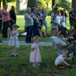 Grupa ludzi stoi w lesie, na trawie. Kobiety, mężczyźni i dzieci obserwują coś. Jasne słońce oświetla tło.