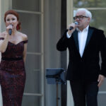 Dwoje ludzi śpiewa na scenie; kobieta w czerwonej sukni, mężczyzna w czarnym garniturze. Tło jasne, kolorowe światła.