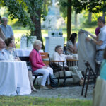 Grupa osób siedzi na zewnątrz, otoczona drzewami. Ubrania w jasnych kolorach; mężczyzna trzyma szarą tkaninę. Stoły przykryte białymi obrusami.