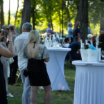 Na zdjęciu ludzie na przyjęciu plenerowym, zielone drzewa w tle. Stoły koktajlowe z białymi obrusami, butelki i kieliszki. Słońce oświetla scenę.