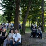 Grupa ludzi siedzi i stoi w parku wśród drzew. Tło to zielone trawniki i las. Scena jest oświetlona naturalnym światłem.
