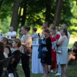 Na zdjęciu jest grupa ludzi na zewnątrz w lesie. Ubrani są kolorowo, w tle widać drzewa i stoliki koktajlowe. Wydarzenie odbywa się w słonecznym dniu.