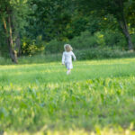 Małe dziecko w kwiecistej sukience biegnie przez zieloną łąkę. Otaczają je wysokie drzewa. Tło jest pełne bujnej roślinności.
