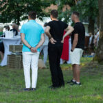 Trzech mężczyzn stoi na trawie, tyłem do kamery. Otoczenie to zielone drzewa i grupa ludzi wokół białych stolików. Jeden mężczyzna ma turkusową koszulkę, reszta jest ubrana w czerń.