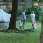 Na zdjęciu dwie osoby dorosłe i dwoje dzieci spacerują po trawniku. W tle ceglana ściana i zieleń drzew. Obok stoi okrągły stół z koszem.