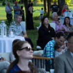 Grupa ludzi siedzi na zewnątrz. Stoły z białymi obrusami i dekoracjami. Tło to zielona przestrzeń z drzewami.