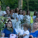 Grupa ludzi na zewnątrz, siedzących przy stołach pod drzewami. Tło jest zielone i naturalne. Dekoracje w bieli i zieleni.