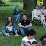 Grupa ludzi siedzi na trawie w parku, w tle drzewa. Większość ubrana w letnie ubrania, dominują zielenie i brązy. Atmosfera jest spokojna i zrelaksowana.