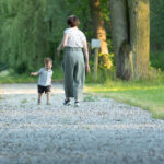 Kobieta i dziecko spacerują po żwirowanej ścieżce w lesie. Zieleń drzew dominuje w tle. Jasne, spokojne otoczenie.