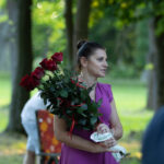 Kobieta w różowej sukience trzyma bukiet czerwonych róż. Tło to zielony las z promieniami słońca. Scena jest letnia i spokojna.