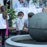 Na zdjęciu widoczna jest grupa ludzi obok fontanny z kulą. Dzieci w kolorowych ubraniach, tło z roślinnością i świecami. Scena toczy się na zewnątrz na tle zieleni i kamienistego podłoża.