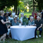 Grupa ludzi siedzi przy białym stole w parku. W tle drzewa i więcej osób. Atmosfera spotkania plenerowego.