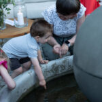 Dzieci bawią się przy fontannie, zanurzając ręce w wodzie. Kobieta w białej bluzce z wzorem serc obserwuje, siedząc obok. Stół z kwiatami i świecą w tle.