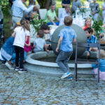 Dzieci bawią się wokół fontanny przy okrągłym stole. Tło to grupa dorosłych i zielona przyroda. Dominuje kolor niebieski i zielony.