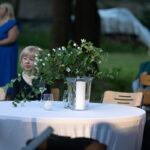 Stół z białym obrusem, na którym stoi szklany wazon z zielonymi roślinami i świecą. W tle osoby siedzą na krzesłach. Sceneria zewnętrzna, zielona trawa.