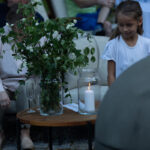 Na drewnianym stoliku stoi wazon z zielonymi gałązkami i szklany lampion z białą świecą. Blisko siedzi osoba w jasnej bluzce i dziewczynka. W tle widać osoby w rozmytych kolorach.