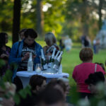 Ludzie stoją przy stoliku z lodem i butelkami, w parku pełnym drzew. Dominuje zieleń tła, stonowane barwy. Atmosfera relaksu na świeżym powietrzu.