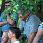 Grupa ludzi na przyjęciu w plenerze, pod drzewem. Kolory zieleni z liści, niebo i ubrania. Mężczyzna w szarej koszulce centralnie.