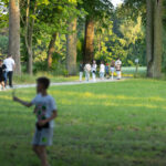 Grupa ludzi spaceruje po leśnej ścieżce, otoczonej drzewami. Dookoła dominuje zielona trawa i drzewa. W tle widać osoby w letnich ubraniach.