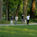 Na zdjęciu widać pięć osób spacerujących po leśnej ścieżce. W tle widać drzewa i soczystą zieleń. Mężczyzna, kobiety i dziecko odwróceni są w kierunku lasu.