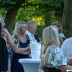 Grupa ludzi rozmawia na świeżym powietrzu przy stoliku. W tle zielone drzewa. Białe i kolorowe ubrania dominują.