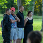 Grupa ludzi stoi na trawie w parku. Mężczyzna w czerwonej czapce sięga pod brodę. Tło to zielone drzewa i trawnik.