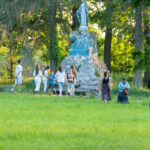Grupa ludzi stoi przy starym, niebieskim pomniku w parku. W tle widać zielone drzewa i trawę. Atmosfera jest spokojna.