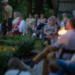 Grupa osób siedzi na zewnątrz przy stołach, wokół roślin. Tło zielone, wieczorne światło dodaje przytulności. Scena jest spokojna i towarzyska.