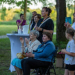Grupa ludzi stoi i siedzi na trawniku, na tle drzew. Kobieta w czerni trzyma kieliszek, obok biały stolik z napojami. W tle jasnozielone pola i drzewa.
