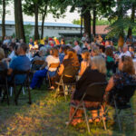 Grupa ludzi siedzi na zewnątrz na krzesłach wśród drzew. Jest wieczór, ciepłe światło oświetla twarze. W tle widoczne są drzewa i budynek.