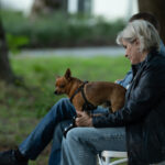 Na zdjęciu dwie osoby siedzą na białych krzesłach w parku. Jedna osoba trzyma brązowego psa na kolanach. W tle rozmyte drzewa i zieleń.