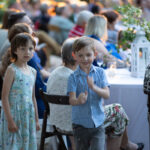 Dwoje dzieci stoi obok stołu podczas przyjęcia. Dziewczynka w kwiecistej sukience, chłopiec w niebieskiej koszuli. Tło jest rozmyte, widać ludzi przy stole.