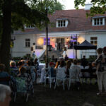 Plenerowy koncert przed białym budynkiem z czerwonym dachem. Publiczność siedzi na krzesłach, scena oświetlona żółtymi i fioletowymi reflektorami. W tle drzewa dodają atmosfery.