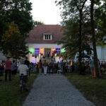 Publiczność słucha koncertu przed białym budynkiem w otoczeniu drzew. Scena oświetlona kolorowymi światłami. Wieczorny klimat.