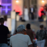 Mężczyzna w czapce na rowerze, patrzący na scenę z ludźmi w tle. Wieczór, kolorowe oświetlenie fioletowe i czerwone. Tłumek obserwuje.