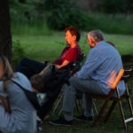 Grupa osób siedzi na krzesłach na trawie w parku. Tło jest zielone z drzewami, a światło zachodzącego słońca oświetla scenę. Dwie z osób intensywnie rozmawiają.