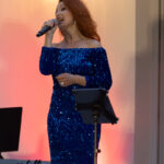 Kobieta w niebieskiej, błyszczącej sukni śpiewa do mikrofonu. Tło jest w odcieniach czerwieni. Widać stojak na nuty i klawiaturę.