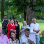 Na zdjęciu grupa ludzi na świeżym powietrzu. Osoby stoją przy stole wśród zieleni drzew. W tle widać zrelaksowaną atmosferę.