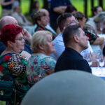 Na zdjęciu widać grupę ludzi przy stole, na zewnątrz na zielonej trawie. Osoby są odwrócone, zwracają uwagę na coś poza kadrem. Kolory są naturalne, dominuje zieleń i różnorodne kolory ubrań.